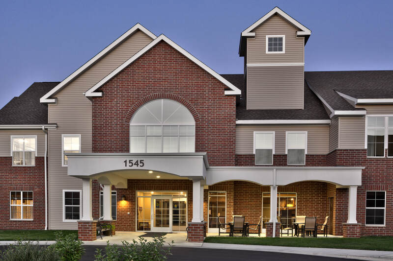 GracePointe Crossing exterior entrance, highlighting brick, siding, columns, ample windows, and outdoor residential seating.   